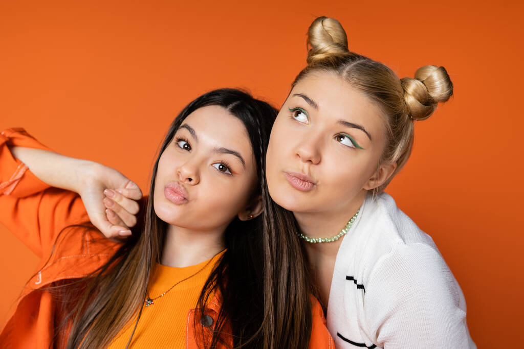 Porträt von stilvollen blonden Teenager-Mädchen mit kühnem Make-up posiert und in der Nähe brünette Freundin schmollen Lippen isoliert auf orange, modische Mädchen mit Sinn für Stil - Foto, Bild