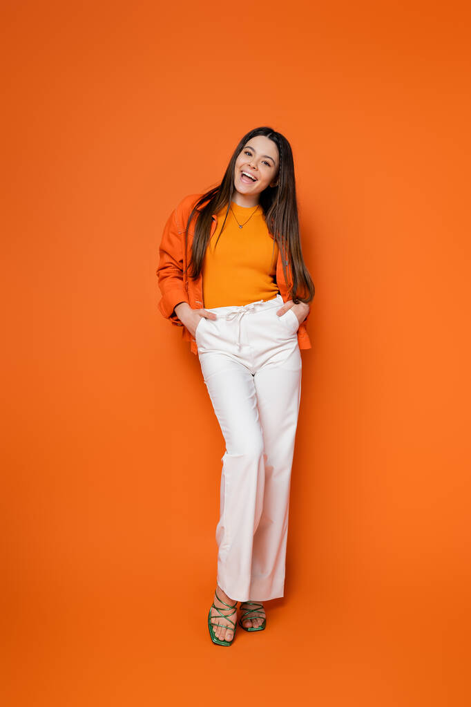 Full length of cheerful and brunette teenage girl in stylish casual outfit holding hands in pockets and looking at camera while standing on orange background, cool and confident teenage girl - Photo, Image