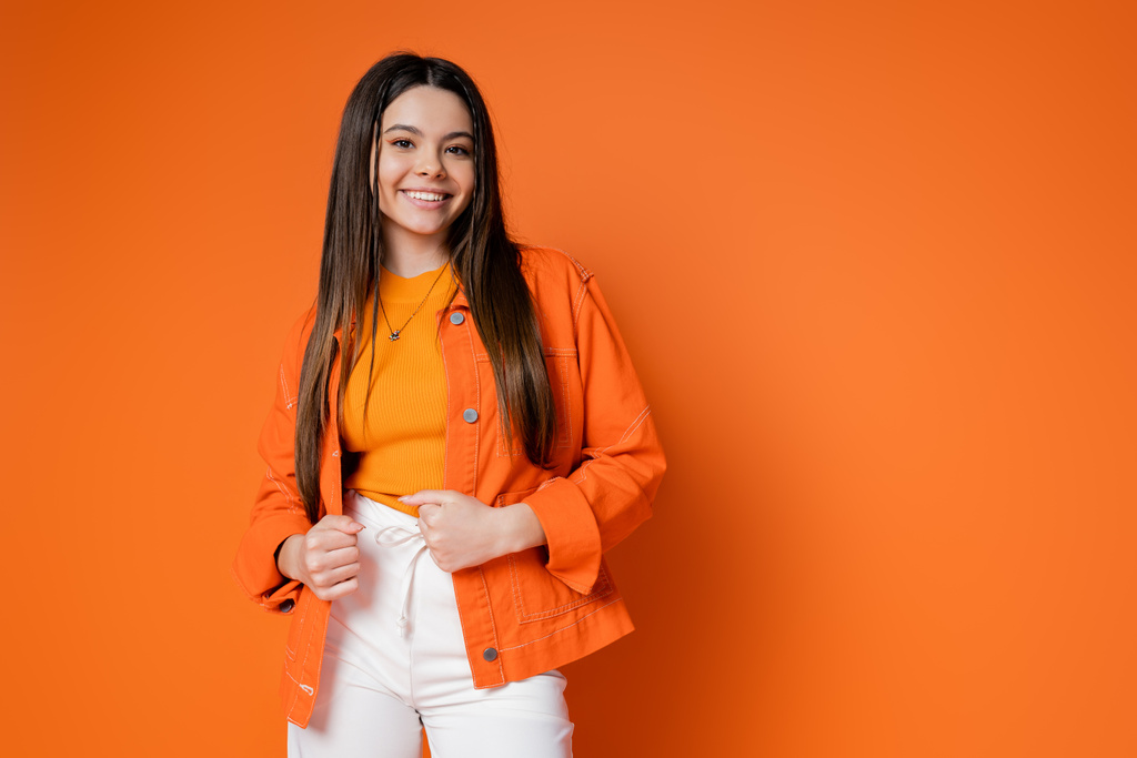 Fashionable and brunette teenage girl in denim jacket and pants smiling while looking at camera and posing on orange background, cool and confident teenage girl - Photo, Image