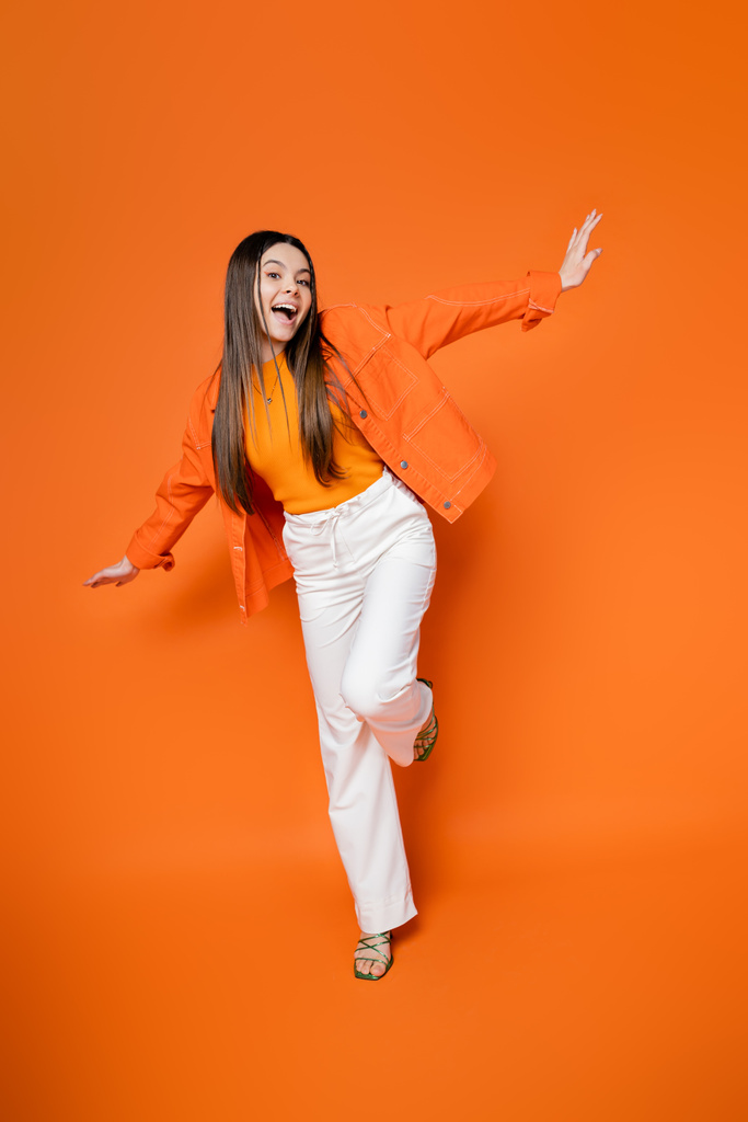 Full length of excited and trendy brunette teen model in denim jacket and pants posing and looking at camera while standing on orange background, cool and confident teenage girl - Photo, Image
