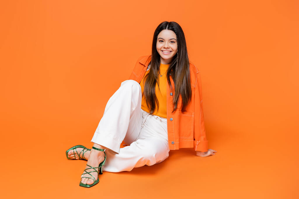 Fashionable and smiling brunette teenage girl in heels and denim jacket looking at camera while sitting and posing on orange background, cool and confident teenage girl, gen z fashion - Photo, Image