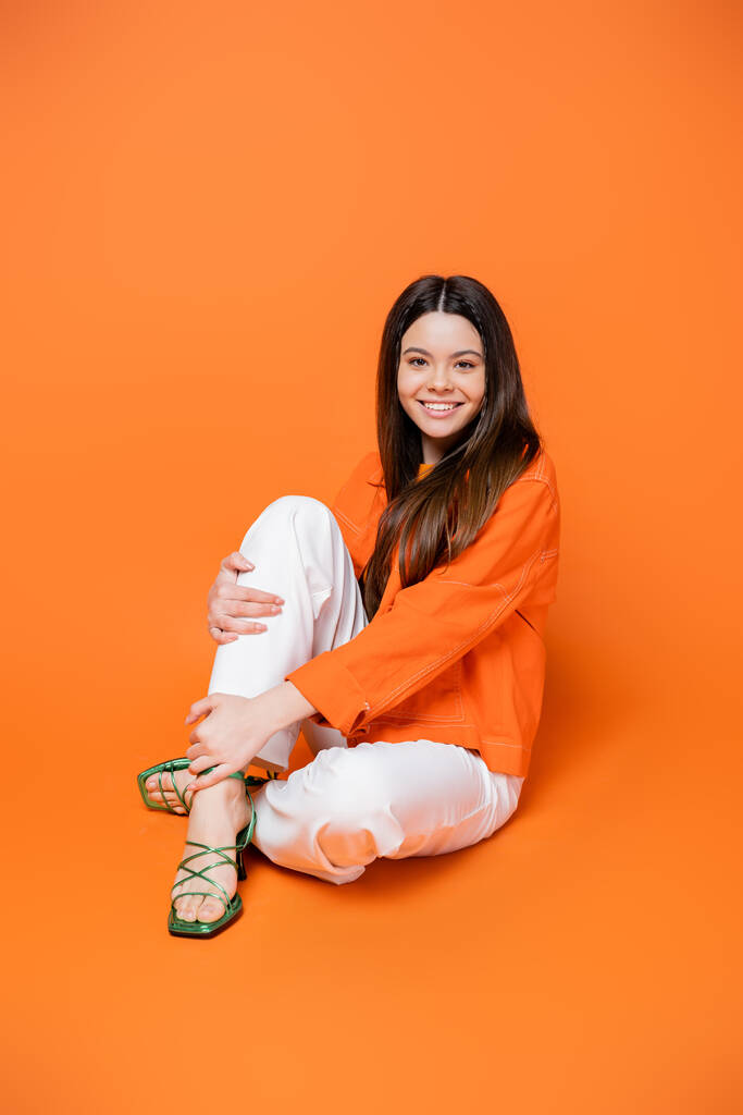 Full length of positive brunette teenager in heels and denim jacket in stylish outfit touching leg while sitting and posing on orange background, cool and confident teenage girl, gen z fashion - Photo, Image