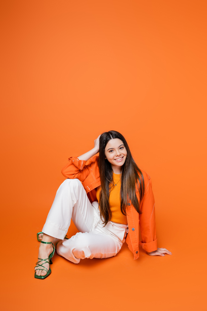 Trendy brunette teenage girl in denim jacket, pants and heels posing and smiling at camera while sitting on orange background, cool and confident teenage girl, gen z fashion - Photo, Image