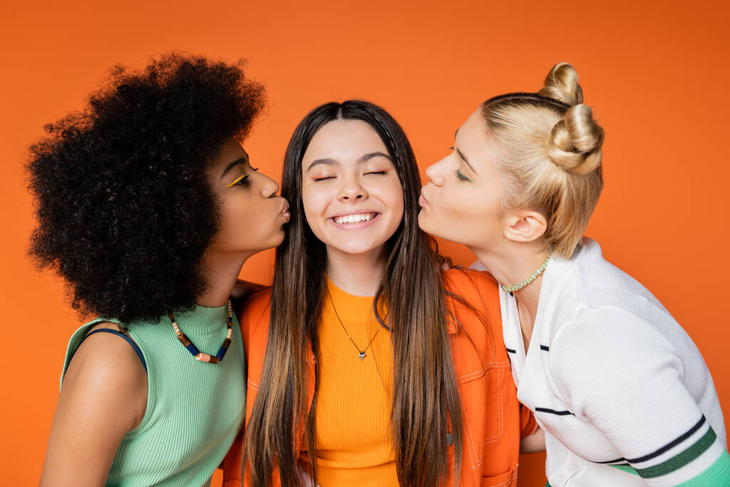 Подростковые африканские американские и блондинистые подружки с красочным макияжем целуются с веселыми и стильными подружками, стоя изолированными на оранжевых, модных нарядах и модных нарядах - Фото, изображение