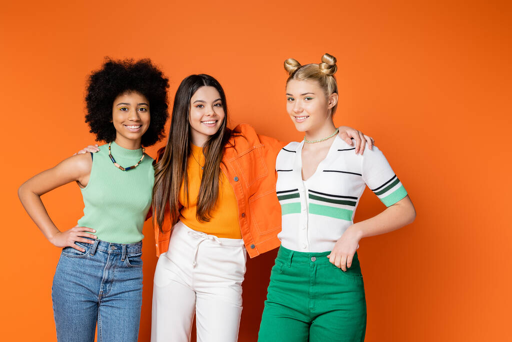 Lächelndes brünettes Mädchen in lässigem Outfit umarmt gemischtrassige Freundinnen mit trendigem, hellem Make-up und posiert auf orangefarbenem Hintergrund, trendigen Outfits und modisch zukunftsweisenden Blicken - Foto, Bild