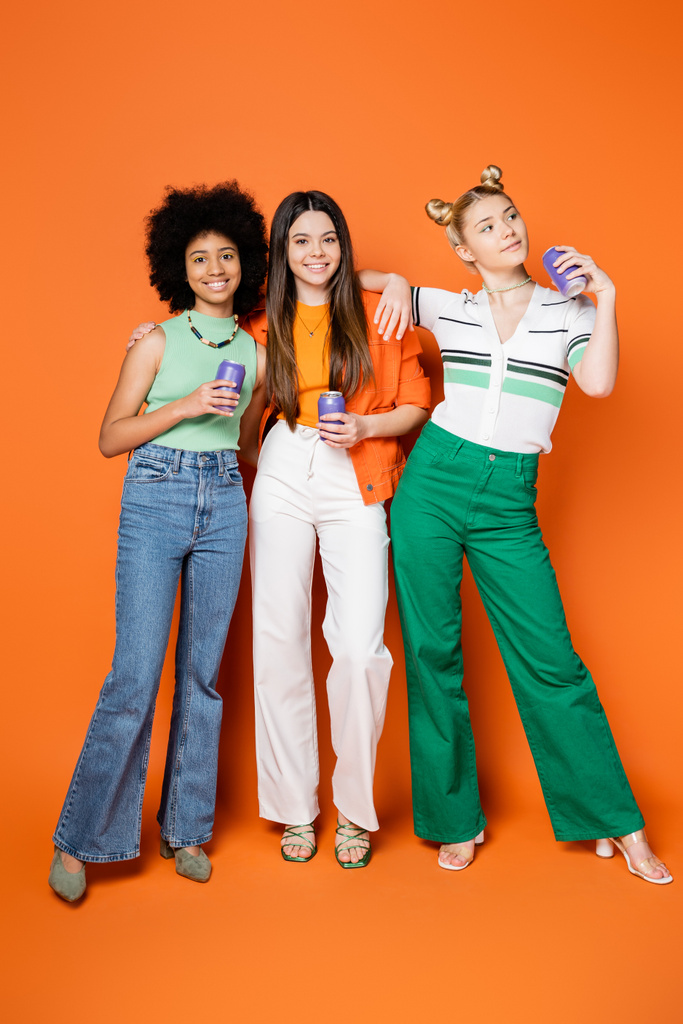 Pełna długość wesołego i stylowego nastolatka przytulając wielonarodowe dziewczyny i trzymając konserwy drinka podczas pozowania razem na pomarańczowym tle, modne stroje i moda-forward wygląd - Zdjęcie, obraz