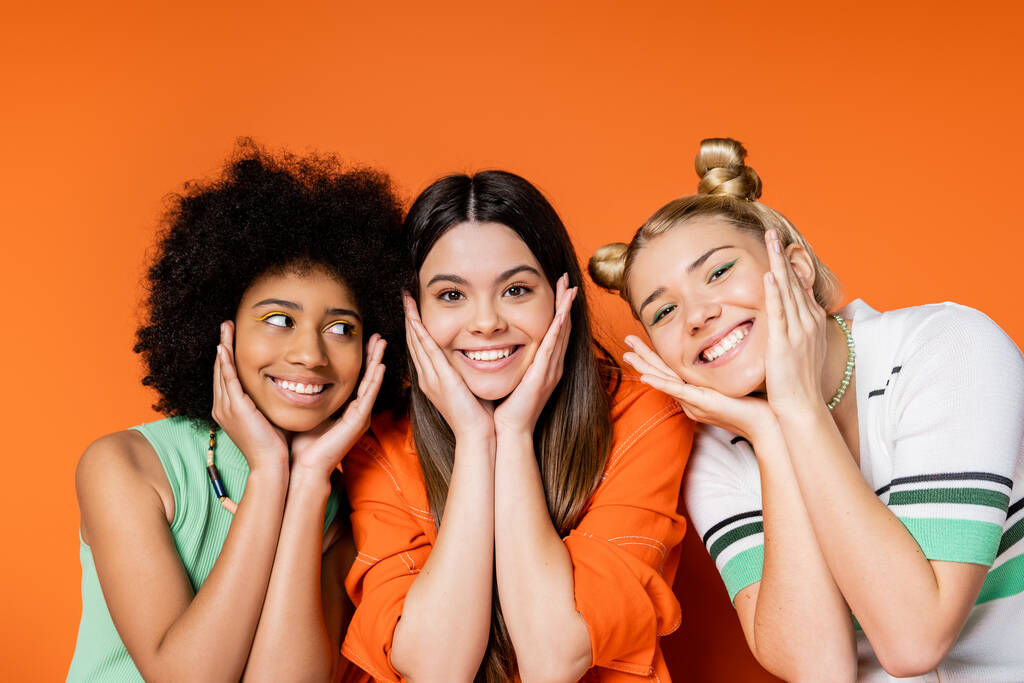 Portret van positieve en interraciale tienervriendinnen met gedurfde make-up en casual kleding die de wangen raken en op een oranje achtergrond staan, trendy outfits en fashion-forward looks - Foto, afbeelding