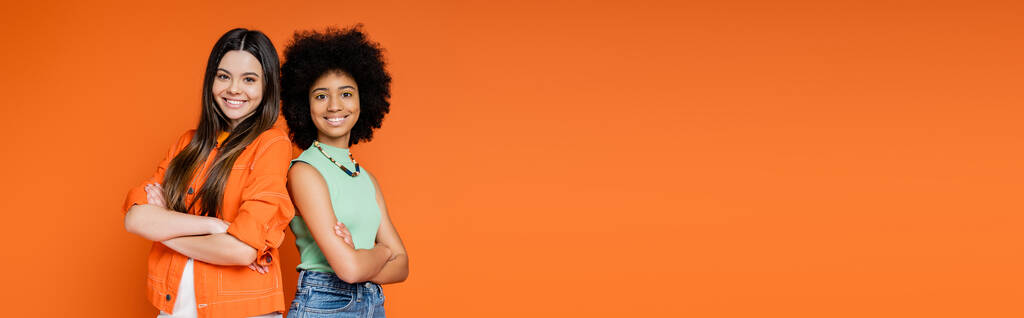 Lachende en zelfverzekerde multi-etnische tiener vriendinnen met gedurfde make-up kruising armen en staan rug aan rug geïsoleerd op oranje, tiener fashionista 's met een onberispelijke stijl concept, banner met kopieerruimte - Foto, afbeelding