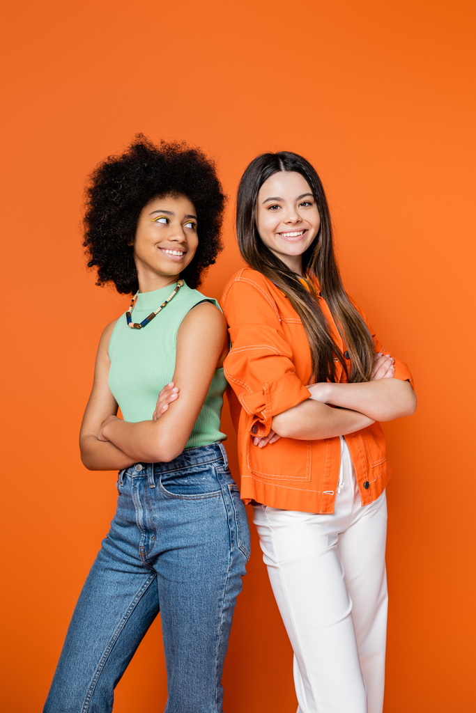 Smiling african american teenage girl with bold makeup crossing arms and standing back to back with stylish girlfriend on orange background, teen fashionistas with impeccable style concept - Photo, Image