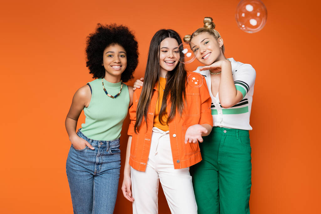 Radosne i wieloetniczne nastolatki w modnych strojach casual pozowanie i stanie w pobliżu baniek mydlanych na pomarańczowym tle, nastolatki mody z nienaganną koncepcją stylu - Zdjęcie, obraz