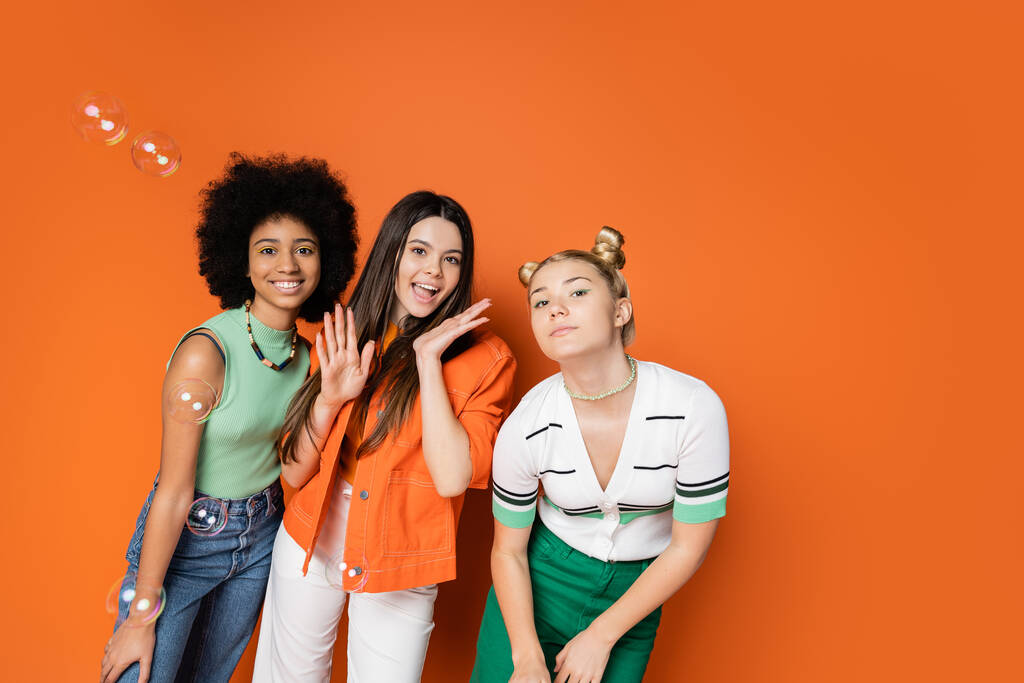 Wieloetniczne i nastolatki dziewczyny w stylowych casual stroje patrząc na aparat i pozowanie w pobliżu baniek mydła stojąc na pomarańczowym tle, nastolatki mody z nienagannym stylu koncepcji - Zdjęcie, obraz