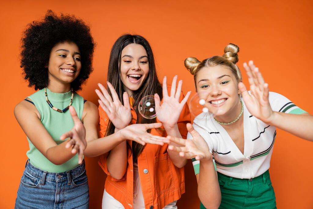 Iloinen ja monikansallinen teini tyttöystäviä tyylikäs rento vaatteita katsomalla saippuakuplia seisoessaan oranssi tausta, teini fashionistas moitteeton tyyli käsite - Valokuva, kuva