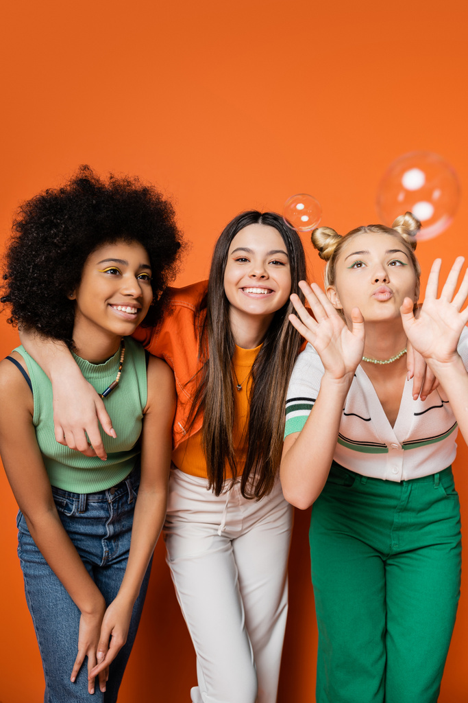 Cheerful brunette teenager hugging stylish multiethnic girlfriends and standing near soap bubbles on orange background, multiethnic teen fashionistas with impeccable style concept - Photo, Image