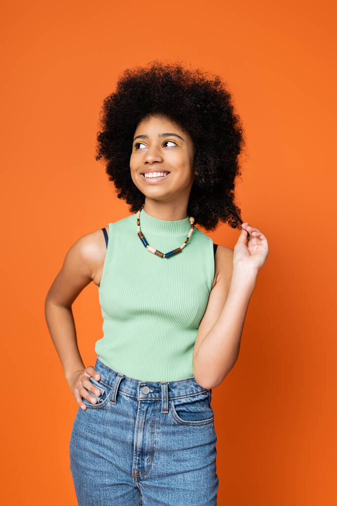 Stylish african american teenage girl in casual outfit and necklace touching hair, holding hand on hip and looking away isolated on orange, trendy teenage girl expressing individuality - Photo, Image