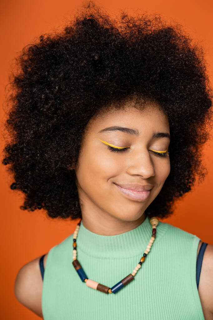 Portrait of joyful and teen african american girl with bold makeup and stylish necklace closing eyes while standing isolated on orange, trendy teenage girl expressing individuality - Photo, Image