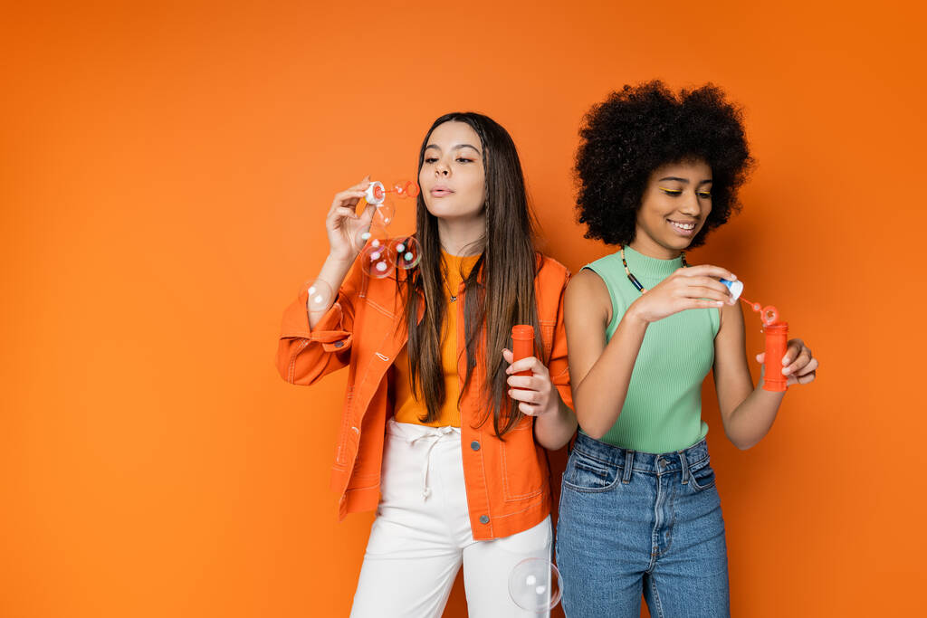 Stylová brunetka dospívající dívka foukání mýdlo bubliny v blízkosti veselý africký americký přítelkyně s tučným make-up zatímco stojí spolu na oranžovém pozadí, módní a módní oblečení - Fotografie, Obrázek