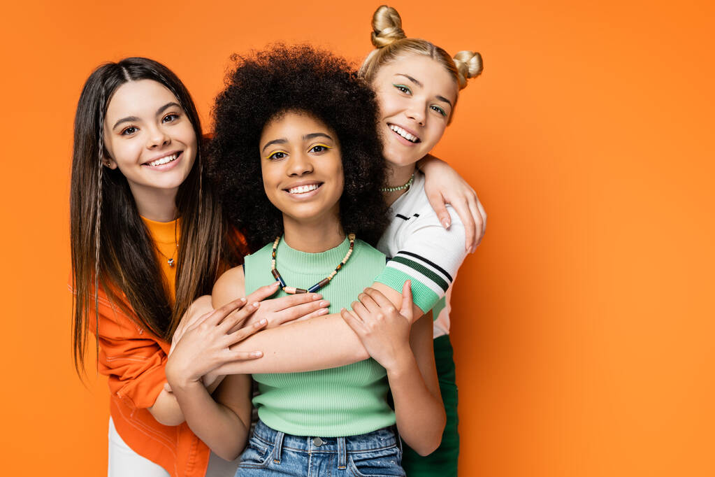 Positieve vriendinnen met kleurrijke make-up knuffelen stijlvolle Afrikaanse Amerikaanse vriendin en samen naar de camera kijken terwijl ze geïsoleerd staan op oranje, modieuze en trendy kleding - Foto, afbeelding