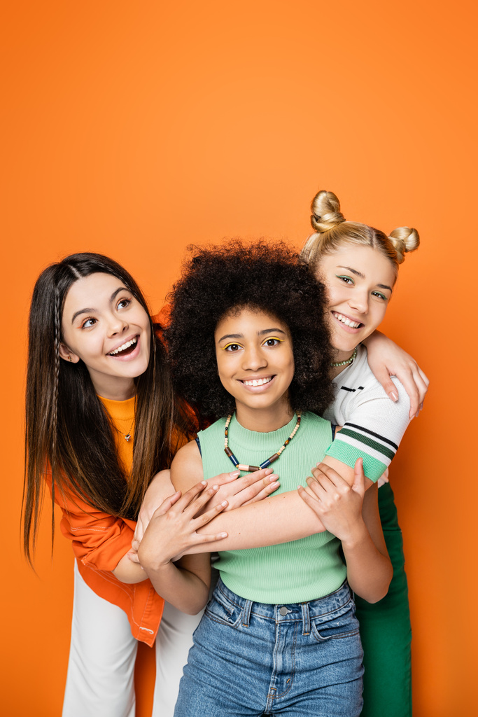 Adolescenti positivi con trucco colorato e abiti casual che abbracciano la ragazza afro-americana insieme e posano su sfondo arancione, vestiti alla moda e alla moda - Foto, immagini