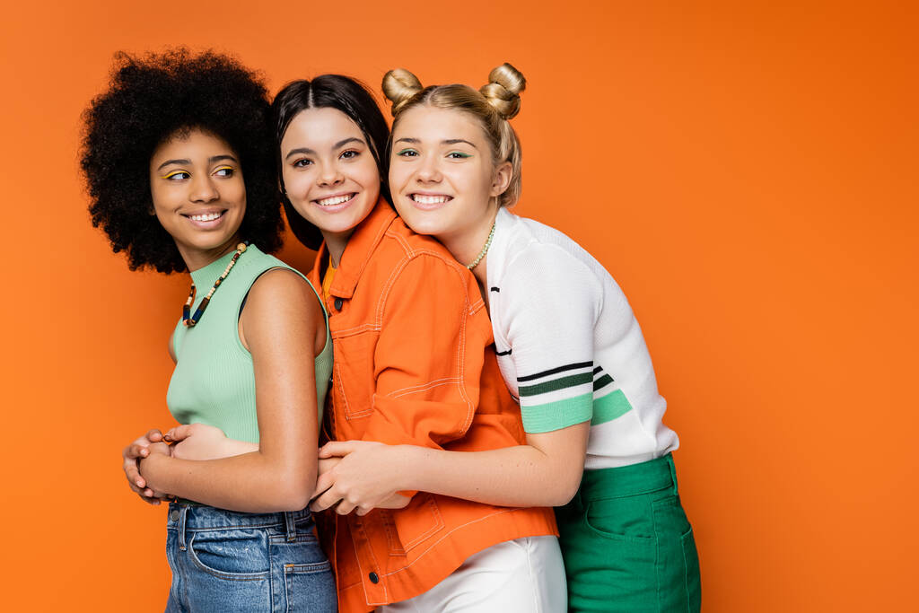Позитивный блондинка подросток с смелым макияжем обнимая стильные многонациональные девушки в повседневных нарядах и глядя на камеру на оранжевом фоне, модная и модная одежда - Фото, изображение