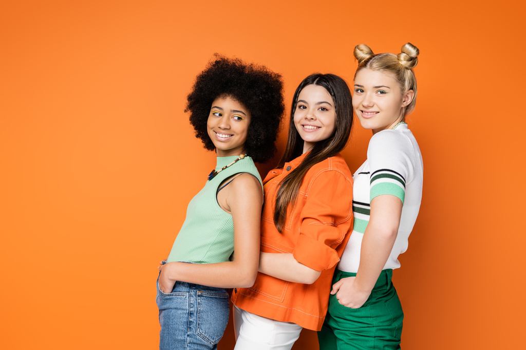 Κομψές και πολυεθνικές έφηβες φίλες με πολύχρωμο μακιγιάζ που κρατιούνται χέρι-χέρι στις τσέπες και ποζάρουν μαζί ενώ στέκονται σε πορτοκαλί φόντο, κομψές και σίγουρες πόζες - Φωτογραφία, εικόνα