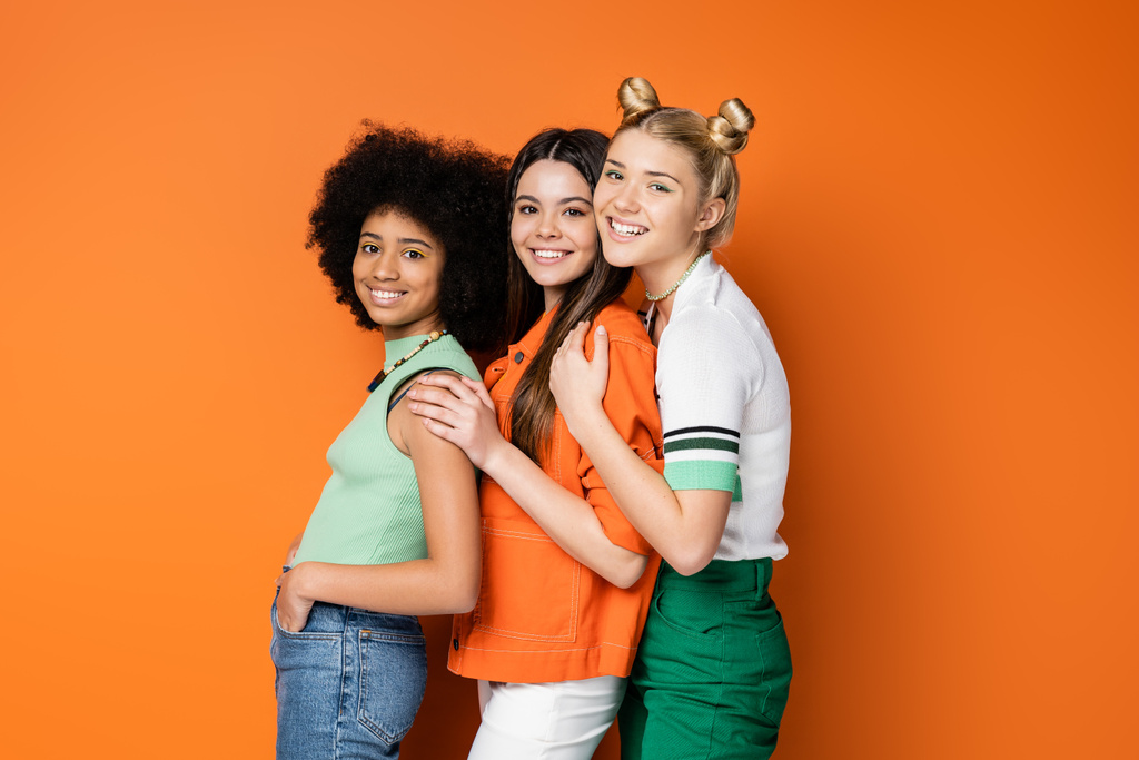 Modne i uśmiechnięte, wielonarodowe nastolatki z odważnym makijażem ubrane w luźne stroje podczas pozowania i patrzenia w kamerę na pomarańczowym tle, stylowe i pewne siebie pozy - Zdjęcie, obraz
