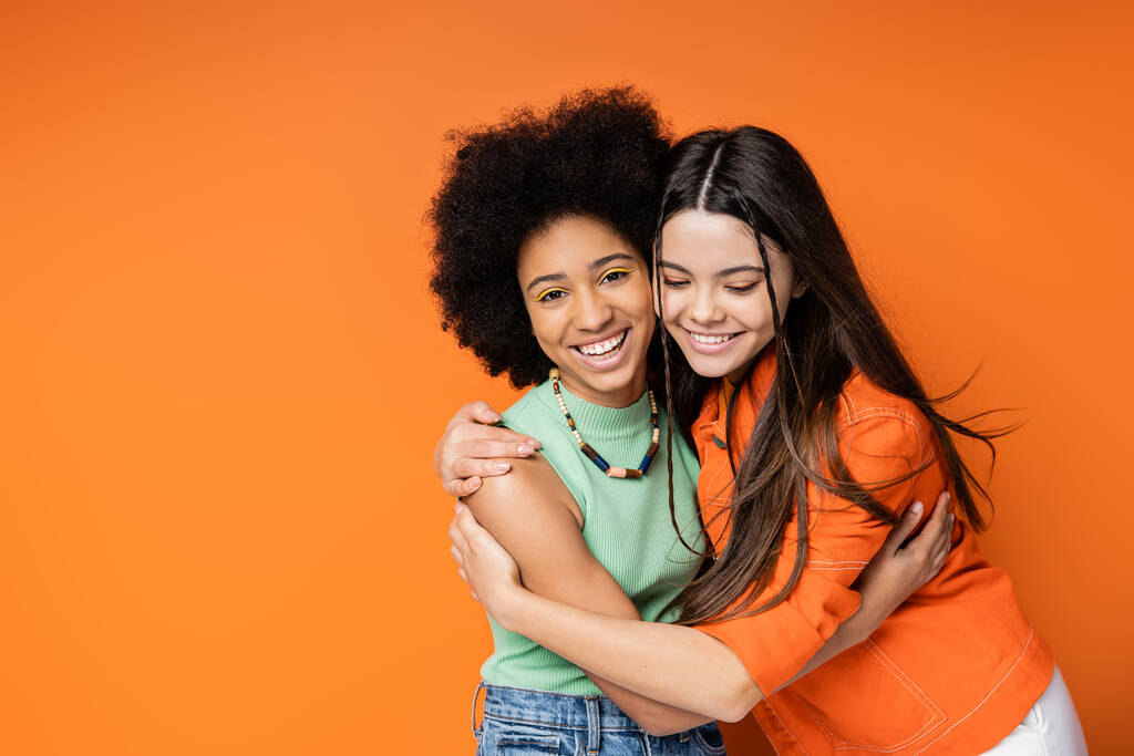 Cheerful and teen african american girl with colorful makeup hugging brunette girlfriend in casual outfit and looking at camera on orange background, stylish and confident poses - Photo, Image