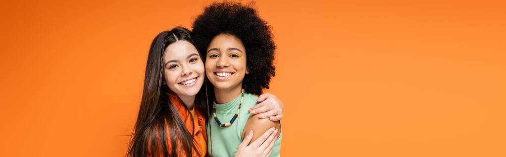 Adolescente brune souriante en vêtements décontractés embrassant une petite amie afro-américaine avec un maquillage audacieux et regardant la caméra isolée sur des poses orange, élégantes et confiantes, bannière  - Photo, image