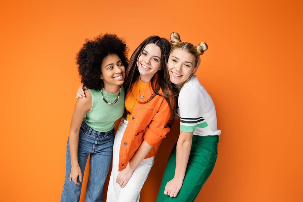 Positive and stylish multiethnic teen girlfriends with colorful makeup hugging brunette friend and posing together on orange background, trendy and stylish hairstyles - Photo, Image