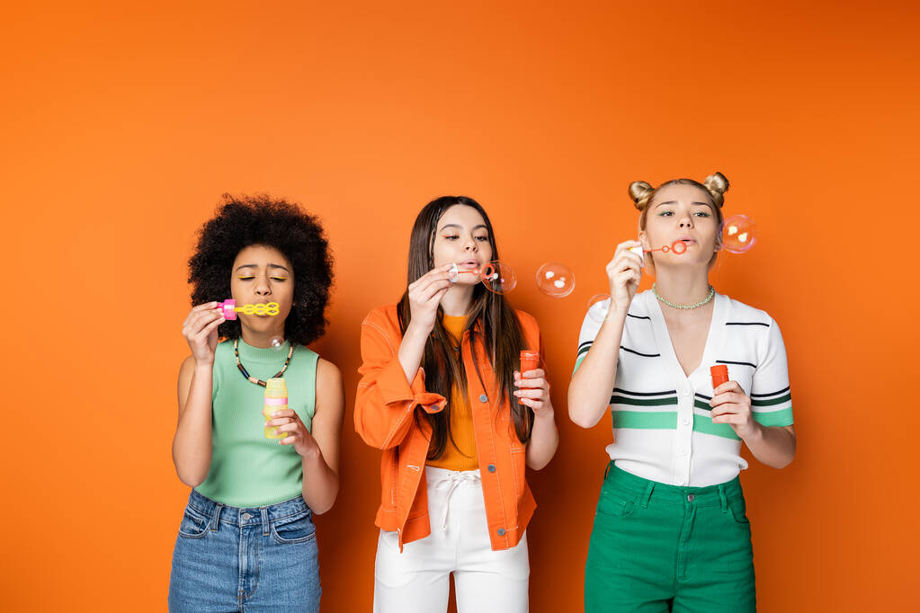 Multiethnic teenage girlfriends with bold makeup and casual outfits blowing soap bubbles and spending time together while standing on orange background, trendy and stylish hairstyles - Photo, Image