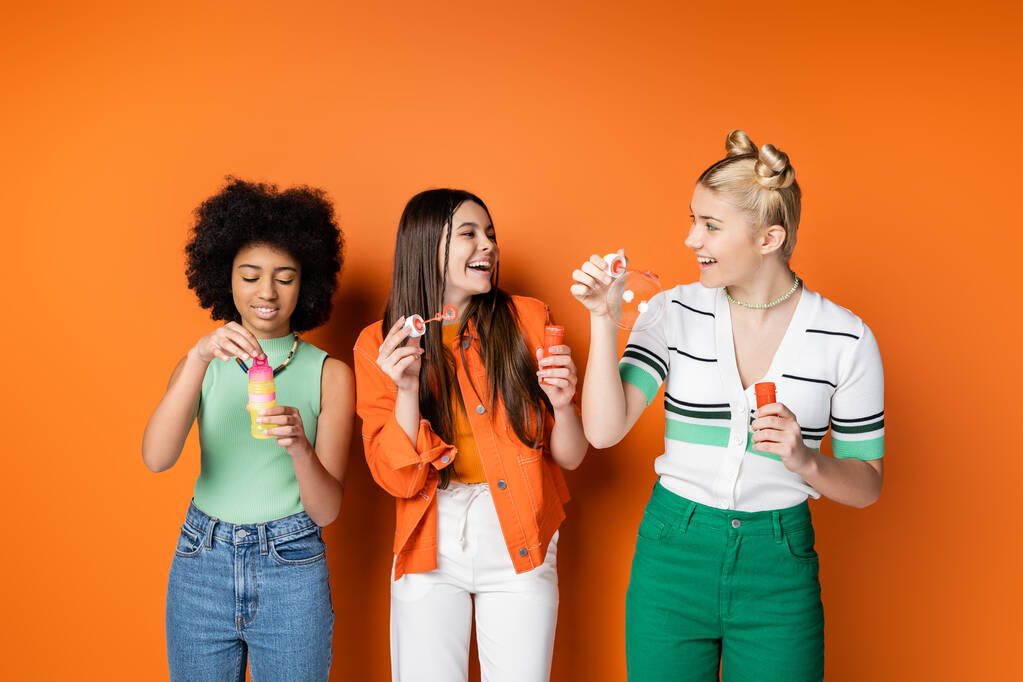 Positive and fashionable teenage girls with colorful makeup wearing casual outfits and holding soap bubbles near african american girlfriend on orange background, trendy and stylish hairstyles - Photo, Image