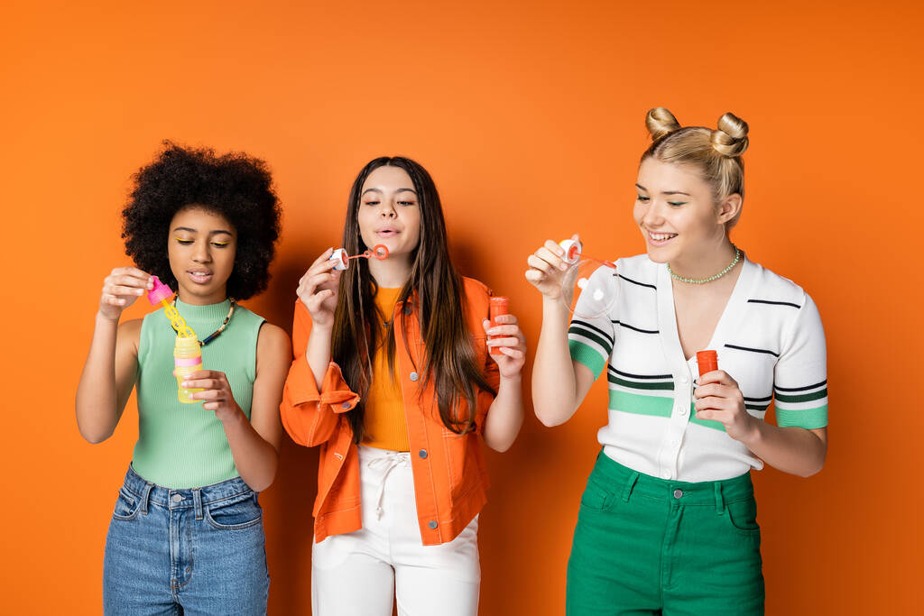 Menina adolescente loira alegre em roupas casuais segurando bolhas de sabão perto de namoradas multiétnicas com maquiagem colorida enquanto está em pé sobre fundo laranja, penteados na moda e elegantes - Foto, Imagem