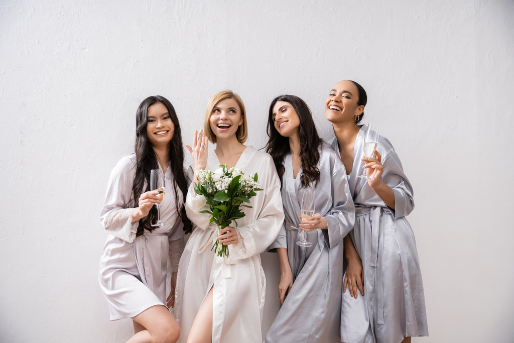 Brautparty, multikulturelle Frauen mit Gläsern mit Champagner, Braut mit weißen Blumen, die ihren Verlobungsring zeigt, Brautjungfern, Vielfalt, Positivität, Brautstrauß, grauer Hintergrund  - Foto, Bild