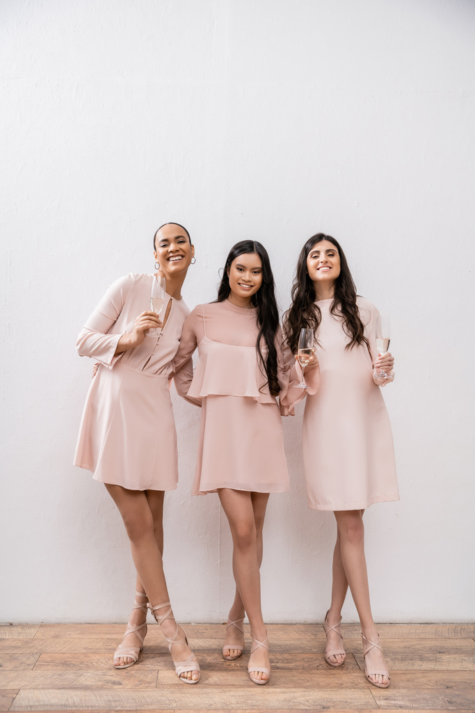 tres damas de honor multiculturales, mujeres bonitas en vestidos de color rosa pastel sosteniendo copas de champán sobre fondo gris, diversidad cultural, moda, celebración, salud  - Foto, Imagen