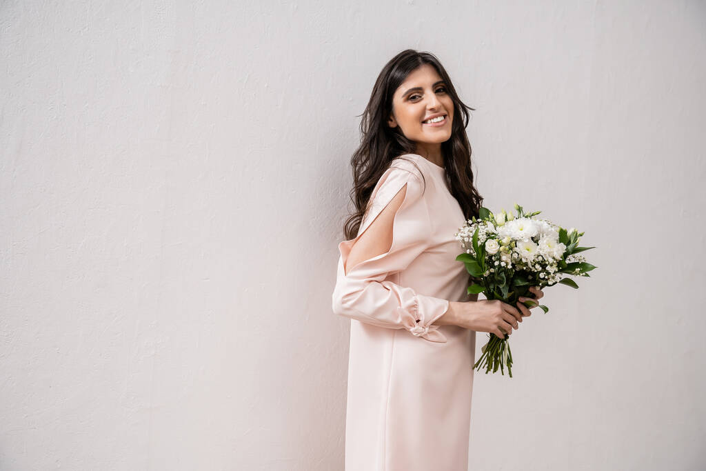 позитивна подружка нареченої в пастельній рожевій сукні, що тримає весільний букет, брюнетка на сірому фоні, білі квіти, особливий випадок, весілля, мода, посмішка і радість, дивлячись на камеру
  - Фото, зображення