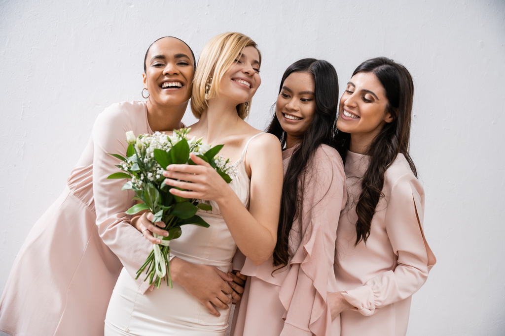 ενθουσιασμός, πολυπολιτισμικές παράνυμφοι αγκαλιάζουν χαρούμενη νύφη με νυφικό μπουκέτο, γκρι φόντο, φυλετική ποικιλομορφία, μόδα, μελαχρινή και ξανθιά, ομάδα γυναικών, λευκά λουλούδια  - Φωτογραφία, εικόνα