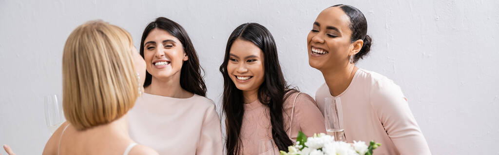 wedding preparations, cheerful interracial bridesmaids looking at blonde bride with bouquet on grey background, admire her style, fitting, bridesmaid gowns, diversity, banner  - Photo, Image
