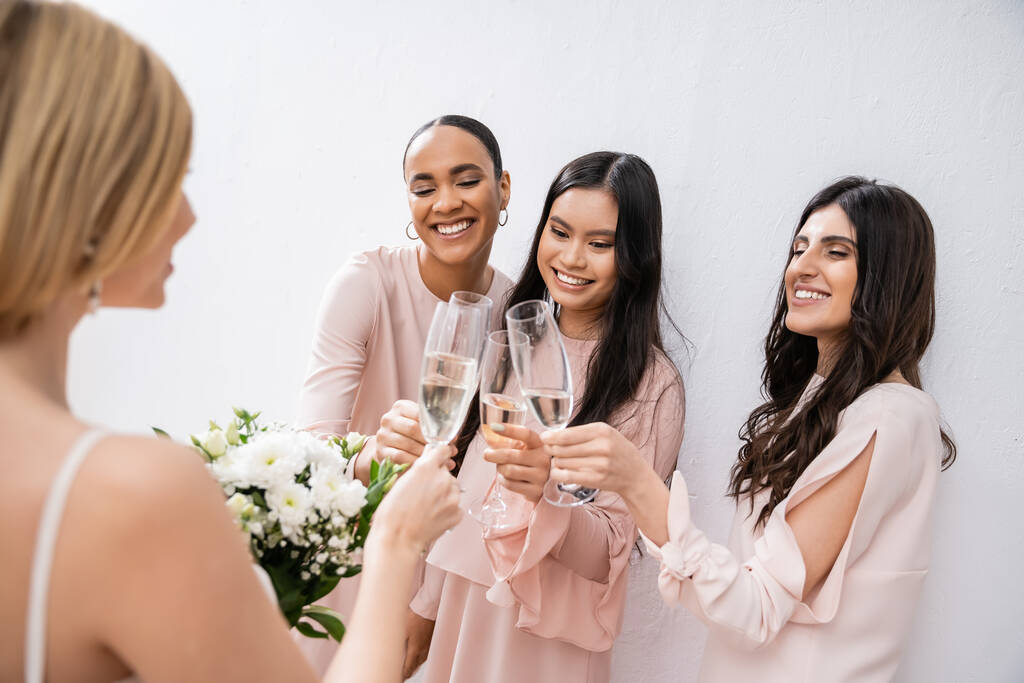 gelukkig interraciale vriendinnen klinkende bril met champagne, bruid met witte bloemen, brunette en blonde vrouwen, bruidsmeisjes, diversiteit, positiviteit, bruidsboeket, grijze achtergrond  - Foto, afbeelding