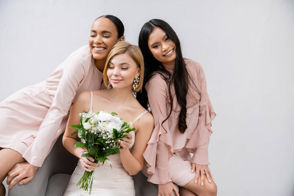 wedding theme, cultural diversity, three women, cheerful bride with bouquet and her interracial bridesmaids sitting on armchair on grey background, brunette and blonde, joy, celebration  - Photo, Image
