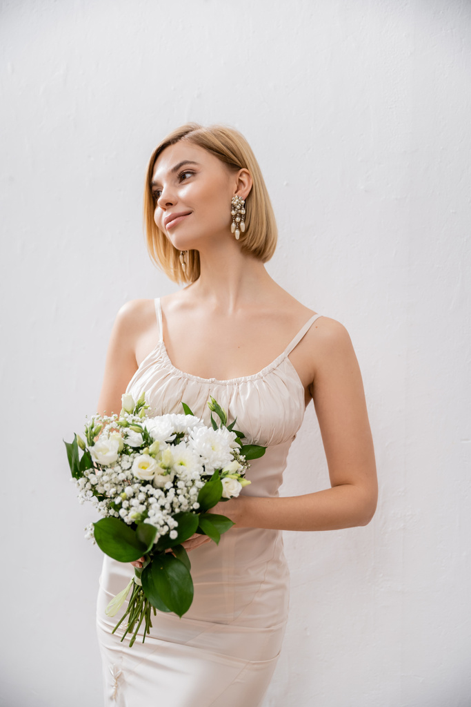 приваблива і блондинка наречена у весільній сукні тримає букет на сірому фоні, білі квіти, весільні аксесуари, щастя, особливий випадок, красива, жіночна, блаженна
  - Фото, зображення