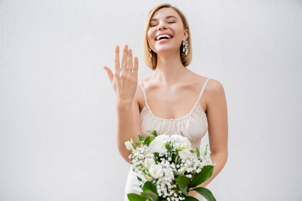különleges alkalom, gyönyörű szőke menyasszony esküvői ruha gazdaság csokor és mutatja eljegyzési gyűrű, fehér virágok, menyasszonyi kiegészítők, boldogság, szürke háttér  - Fotó, kép