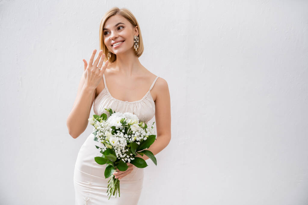 γαμήλιο θέμα, πανέμορφη ξανθιά νύφη σε νυφικό κρατώντας μπουκέτο και δείχνοντας δαχτυλίδι αρραβώνων, λευκά λουλούδια, νυφικά αξεσουάρ, ευτυχία, γκρι φόντο, ειδική περίσταση   - Φωτογραφία, εικόνα