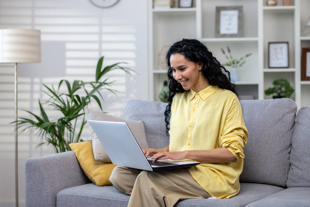 Junge Frau mit Laptop zu Hause auf Sofa sitzend, erfolgreiche Lateinamerikanerin im Wohnzimmer tippt auf Computertastatur, die aus der Ferne online arbeitet. - Foto, Bild