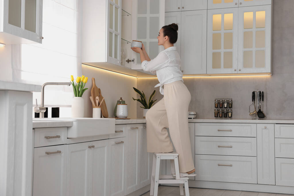 Nainen tikkailla laittamassa valkoista purkkia kaappiin kotona - Valokuva, kuva