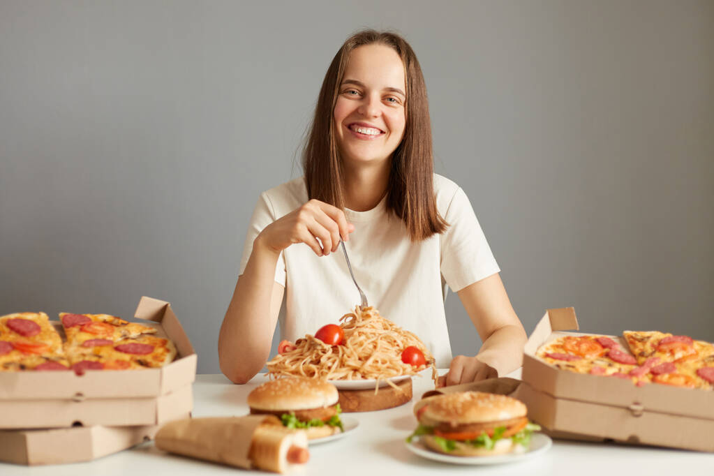 Mujer hermosa sonriente extremadamente feliz con cabello castaño usando camiseta blanca sentada a la mesa entre comida rápida aislada sobre fondo gris disfrutando de una nutrición poco saludable. - Foto, Imagen