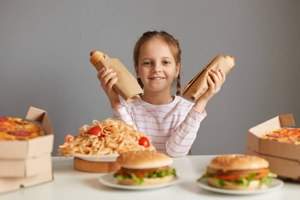 Satisfied delighted little girl with braids sitting at table with fast food isolated over gray background holding two hot dogs looking at cmaera with happy expression. - Photo, Image