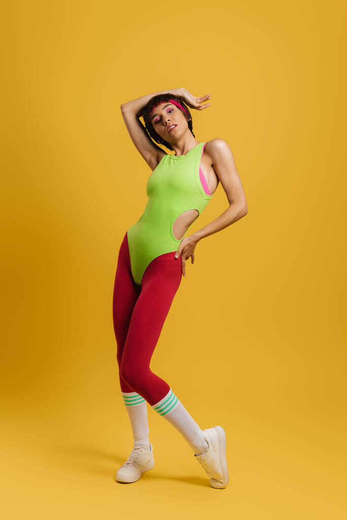 黄色の背景に立ってレトロなスタイルのスポーツウェアでフィット女性の完全な長さ - 写真・画像