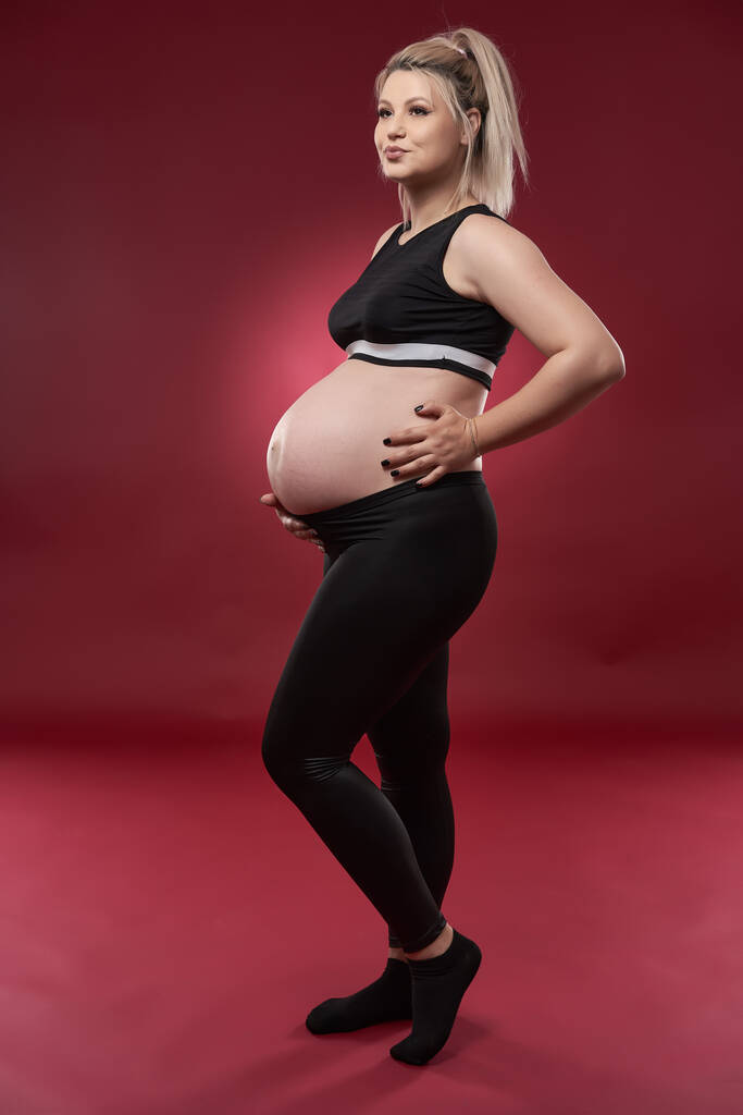 Hermosa embarazada joven rubia en ropa negra, estudio de tiro sobre fondo rojo    - Foto, imagen
