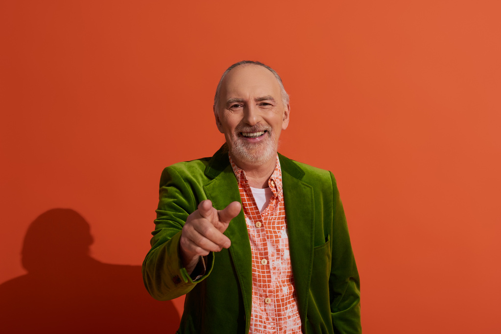 heureux vieillissement, sourire radieux, homme âgé joyeux avec les cheveux gris et la barbe, en chemise à la mode et veste en velours vert regardant la caméra et pointant du doigt sur fond rouge orange - Photo, image