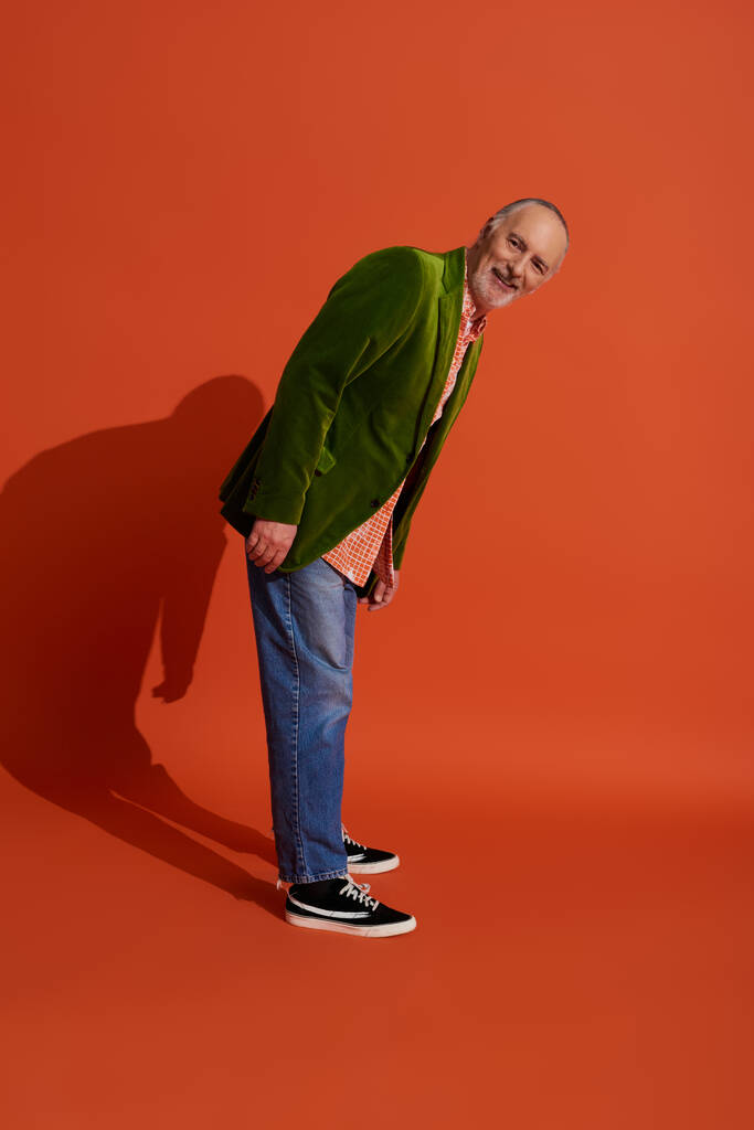 полная длина радостный и бородатый пожилой человек в модной повседневной одежде стоя и глядя на камеру на красном оранжевом фоне, зеленый велюр блейзер, джинсы синие джинсы джинсы, счастливое старение концепции - Фото, изображение