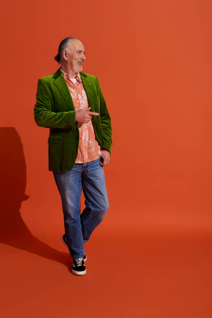 piena lunghezza di uomo anziano fresco e allegro in giacca di velluto verde in piedi con mano in tasca di jeans di jeans blu, guardando altrove e puntando con il dito su sfondo arancione rosso con ombra - Foto, immagini